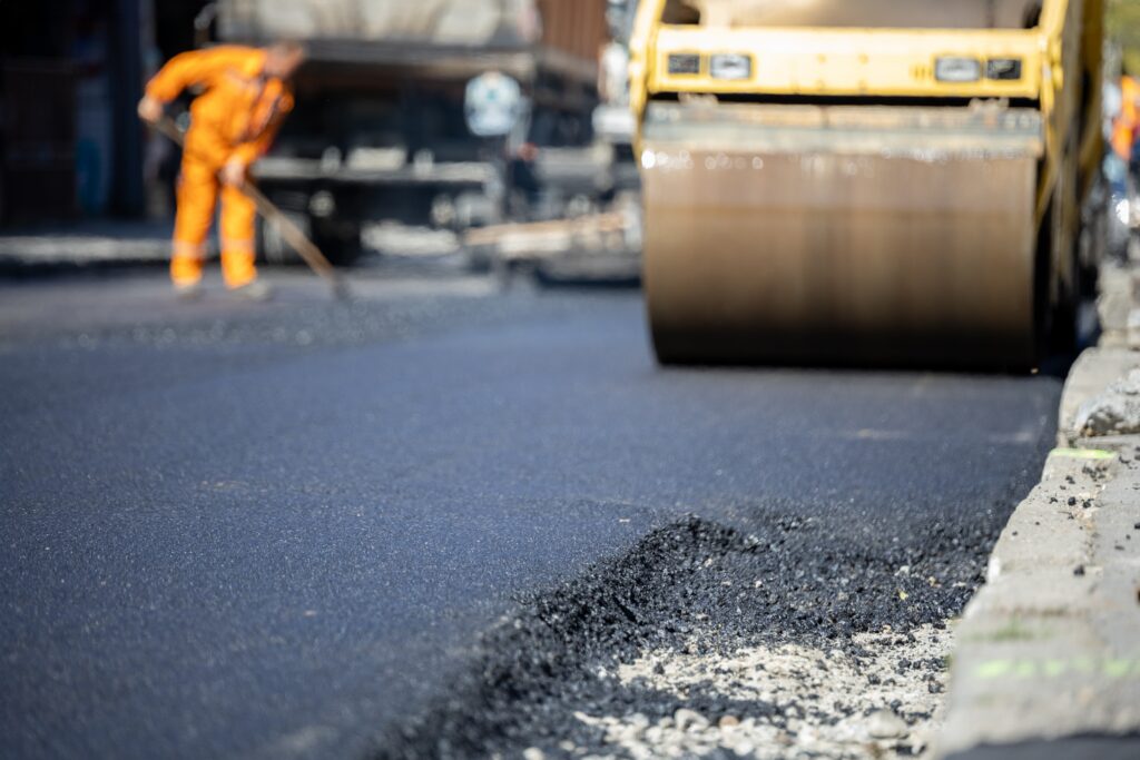 Steamroller smoothing fresh asphalt with worker in orange uniform raking in the background.