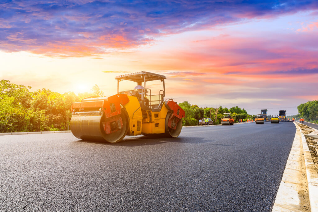 Steamroller paving a new asphalt road at sunset with vibrant sky in the background.