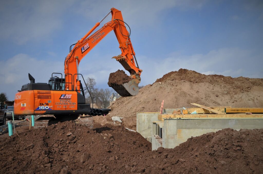 Orange excavator moving dirt at a construction site with soil piles and concrete foundation.