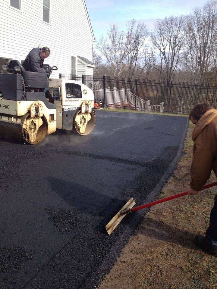Worker operates a roller on freshly laid asphalt, another smooths edges with a rake near a white house and black fence.