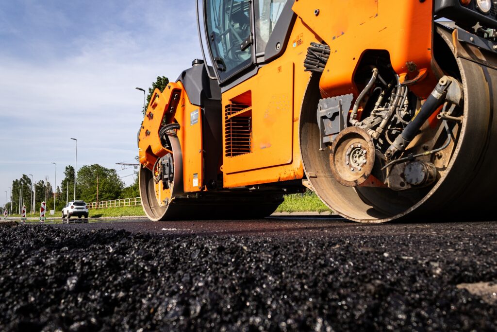 Orange road roller compacting fresh asphalt on a sunny day, with a car and trees in the background.