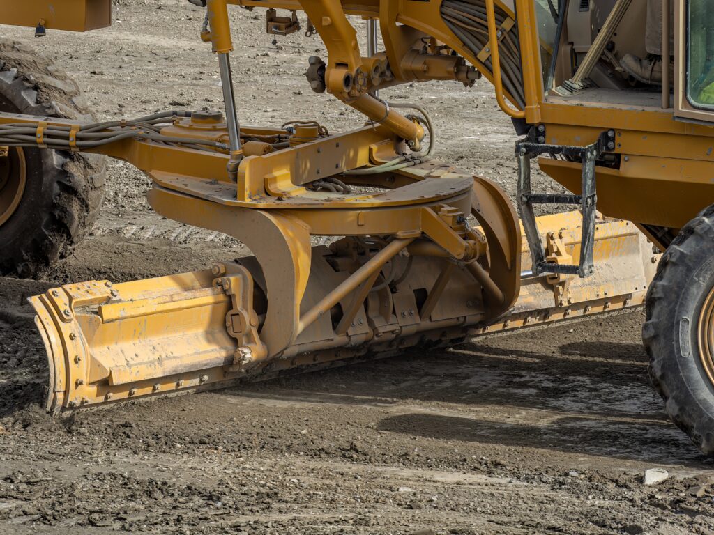 Yellow construction grader smoothing dirt surface on a worksite.